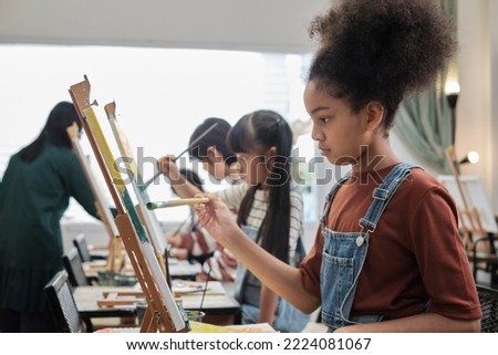 African American girl concentrates on acrylic color picture painting on canvas with students group in art classroom, creative learning with talents and skills in elementary school studio education.