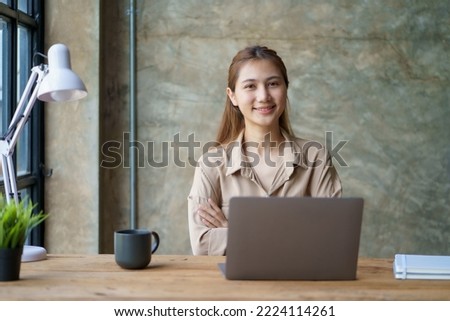 Beautiful Asian businesswoman sitting smiling, cross arms and opened her laptop and happily looking at the camera in the office.