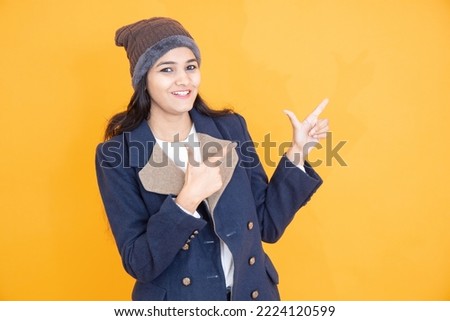 Happy young Indian girl wearing winter cloths pointing at blank space, isolated on orange yellow background, Woman look here pose, Advertisement and promotion.