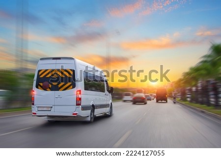 Unrecognizable white small passenger van hurry up on highway at city street traffic with urban cityscape, sunrise sky on background. Charter fast bus van hurry up on with motion blur effect.
