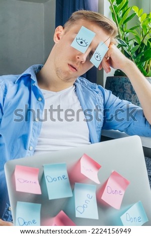 Exhausted businessman with painted eyes on stickers, adhesive notes on face sleeping at home office workplace.Unproductive tired lazy young male dozing working on a difficult project. Funny moment Royalty-Free Stock Photo #2224156949