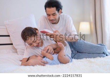 Mom happy feed milk to asian infant baby new born on bed father use mobile phone taking picture