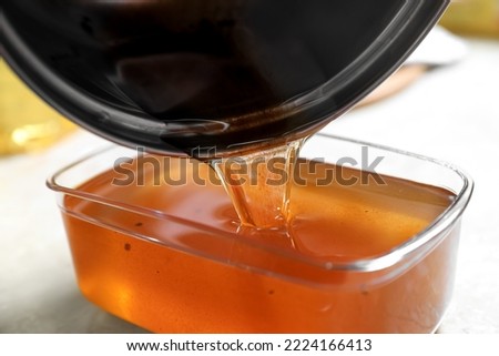 Pouring used cooking oil from saucepan into container on beige table, closeup