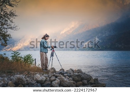 Adult young woman traveler professional photographer taking a picture of the landscape while standing with a camera on a tripod on mountain lake