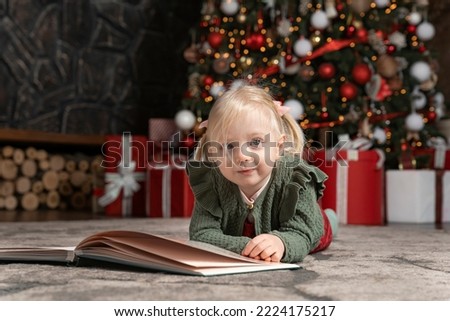 Portrait of cute little blonde girl lying on floor next to large book on Christmas tree background. Children and New Year mood
