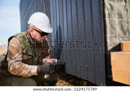 Male builder installing black corrugated iron sheet used as facade of future cottage. Man worker building wooden frame house. Carpentry and construction concept. Royalty-Free Stock Photo #2224175789