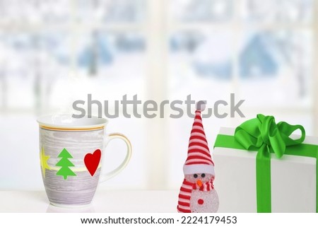Merry Christmas and Happy New Year greeting card. A snowman, a gift package and a Christmas cup on the table in front of abstract winter landscape. Space for text.