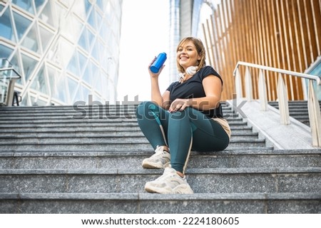 Plus size woman having a break during her workout, curvy young woman in sportswear in cardiovascular training sitting on steps and drinking water, fitness in the city lifestyle, copy space Royalty-Free Stock Photo #2224180605