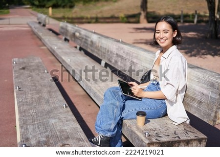 Portrait of asian girl sits on bench in park, draw on her digital tablet with pen, smiling happily, getting creative.