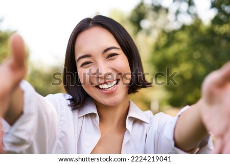 Smiling asian girl takes selfie, video chats, holds smartphone and speaks at camera, posing in park.