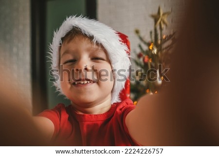 Funny child in Christmas hat takes selfie, the Christmas tree on background. Christmas concept.