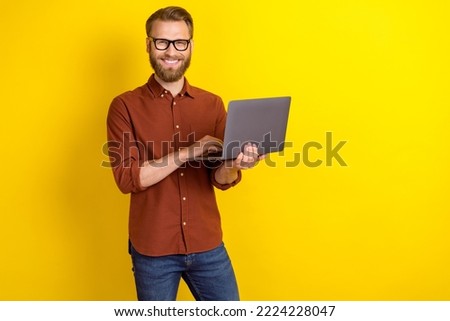 Portrait of good mood toothy beaming man with masculine beard dressed burgundy shirt hold laptop isolated on yellow color background