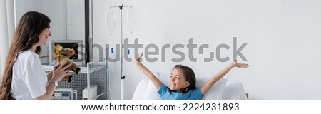 Parent holding soft toy near cheerful daughter in patient gown in clinic, banner