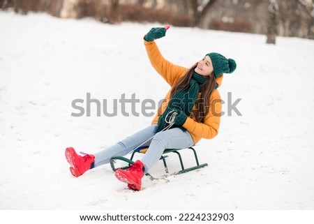 Portrait of beautiful cheerful pre-teen girl wearing warm clothes sitting on sledge taking taking selfie outdoors