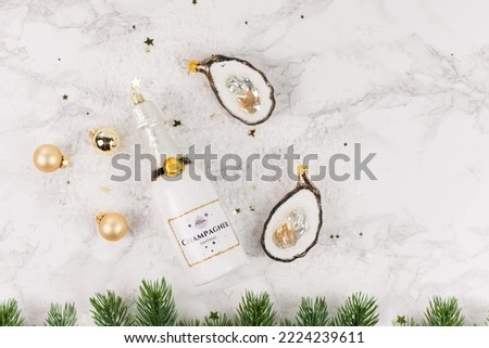 Christmas tree baubles in the shape of a champagne bottle or oysters lie on a marble table. Glitter, stars and fir branches decorate the picture.