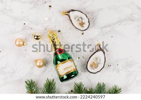 Christmas tree baubles in the shape of a green champagne bottle or oysters lie on a marble table. Glitter, stars and fir branches decorate the picture.