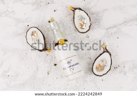 Christmas tree baubles in the shapes of a white champagne bottle or oysters are lying on a marble table. Glitter and golden stars decorate the picture.