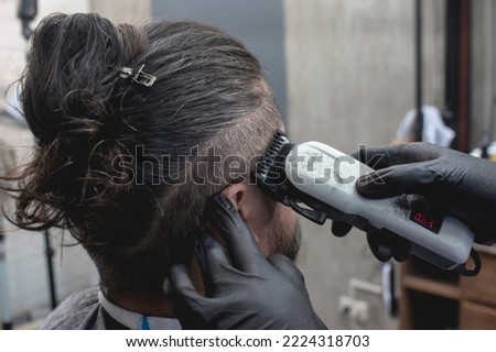 A barber with black gloves uses a white hair clipper to trim the side scalp of a customer. Long hair man bun with fade haircut at a barbershop. Royalty-Free Stock Photo #2224318703