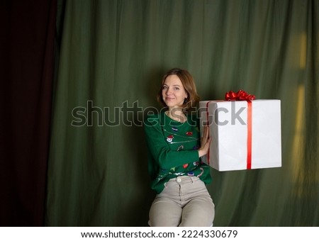 Happy woman 35-40 years old holding a large white gift box tied with a red bow on a dark green background. Surprise, gift, joy. birthday, holiday. Copy space