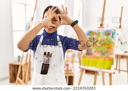 Young brunette woman at art studio doing heart shape with hand and fingers smiling looking through sign 