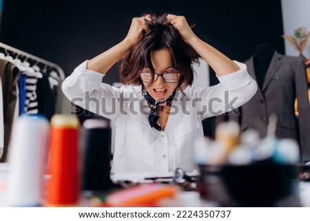 Furious caucasian woman shouting and grabbing dark hair while sitting at desk with sewing equipment around. Angry fashion design lost inspiration and creativity.