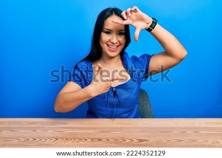 Beautiful hispanic woman with nose piercing sitting on the table smiling making frame with hands and fingers with happy face. creativity and photography concept. 