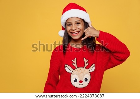 Merry little kid teen girl 13-14 years old wear red xmas sweater Santa hat posing do phone hand gesture like say call me back isolated on plain yellow background Happy New Year 2023 holiday concept