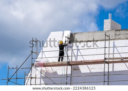Modern construction and insulation of a new residential building. Man installing styrofoam sheets on house facade wall for thermal protection. Worker in protective helmet standing on scaffolding Royalty-Free Stock Photo #2224386123