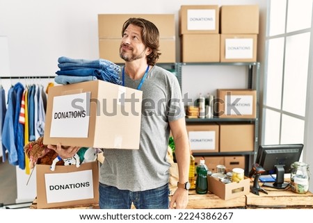 Handsome middle age man holding donations box for charity at volunteer stand smiling looking to the side and staring away thinking. 
