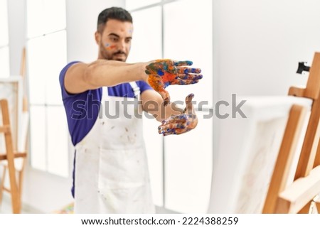 Young hispanic man looking canvas draw doing photo gesture with hands at art studio