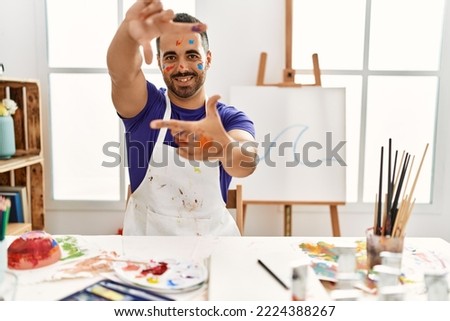 Young hispanic man with beard at art studio with painted face smiling making frame with hands and fingers with happy face. creativity and photography concept. 