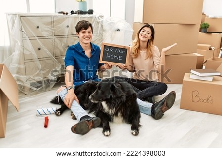 Young caucasian couple with dog holding our first home blackboard at new house smiling cheerful presenting and pointing with palm of hand looking at the camera. 