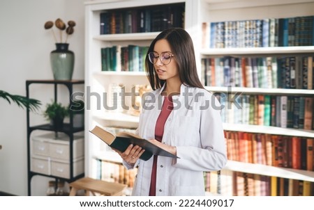 Young concentrated female intern in glasses and white uniform studying medical book while standing near bookcase in modern hospital office Royalty-Free Stock Photo #2224409017