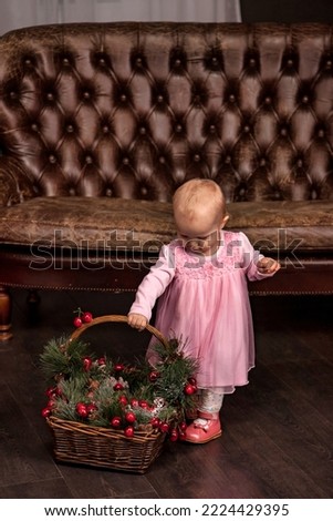 Little girl dressed pink dress with basket Christmas decorated at home, looking down. Baby at Xmas home decorations indoors, background. Concept of cozy home New Year celebration. Copy text space