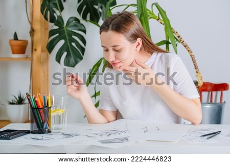 A young woman of European appearance draws with a pencil on white paper. Graphics. The process of drawing with a pencil at home at a white table.