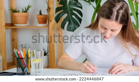 A young woman of European appearance draws with a pencil on white paper. Graphics. The process of drawing with a pencil at home at a white table.