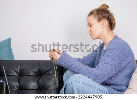 A young reaper knits a hat. The woman is dressed in a sweater in the color of peri peri. The process of knitting a hat on knitting needles. Close angle