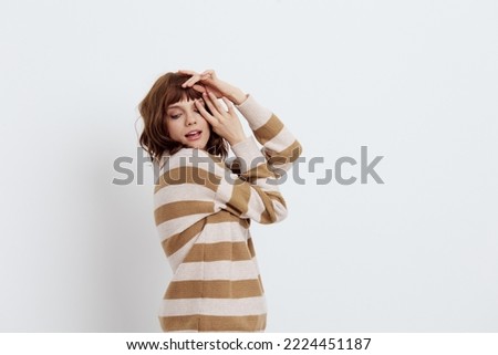 a beautiful, sweet, attractive woman stands in a striped sweater on a light background and raises her hands to her face folded in a relaxed pose. Horizontal photo with empty space