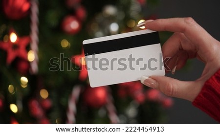Hands of a woman in a red sweater with a credit card on the background of a Christmas tree close-up. Christmas and New Year shopping online, credit card payment. Buying gifts online