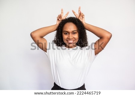 Portrait of happy young woman making bunny ears over white background. African American lady wearing white T-shirt looking at camera and smiling. Fun concept