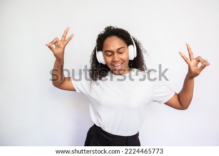 Portrait of happy young woman dancing with closed eyes. African American woman wearing white T-shirt listening to music in headphones over white background. Leisure and music concept