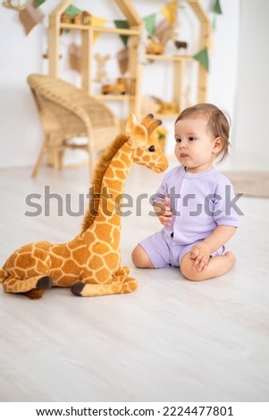 cute smiling healthy baby girl in a purple bodysuit is playing in the nursery at home, looking at a big stuffed giraffe