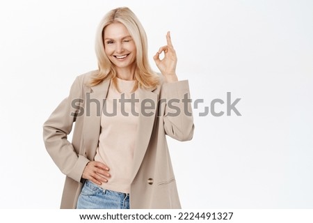 Elegant older woman, blond female model in her 50s, smiling and showing okay, ok sign, wink at you, stands over white background.