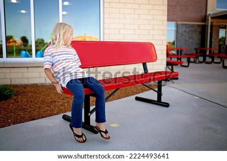 A lonely little girl is sitting on a buddy bench at school waiting for a friend to come play with her on the playground. Royalty-Free Stock Photo #2224493641