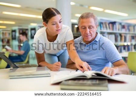 Positive young lady explaining information for mature man while he is using a computer in the library. High quality photo