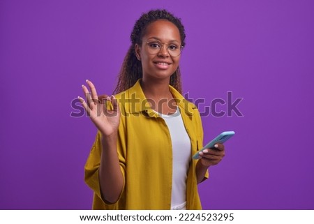 Young successful positive African American woman millennial with smartphone in hand makes OK sign with fingers confirming quality of new phone stands posing on lilac background. OKAY gesture