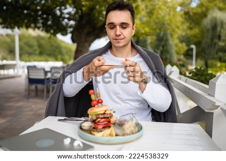 one man caucasian male sitting at terrace of the restaurant using mobile phone to take photo of food burger in a plate on the table in day outdoor real people copy space
