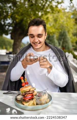 one man caucasian male sitting at terrace of the restaurant using mobile phone to take photo of food burger in a plate on the table in day outdoor real people copy space happy smile