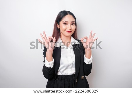 Excited Asian business woman wearing a black suit giving an OK hand gesture isolated by a white background
