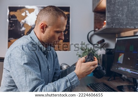 Side view of focused male video editor looking at footage on photo camera while doing post production montage at desktop computer with professional software 